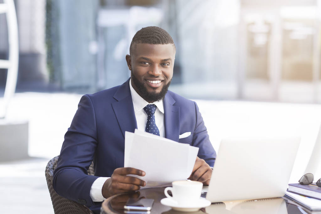 Black Businessman Working With Papers And Laptop In Outdoor Cafe